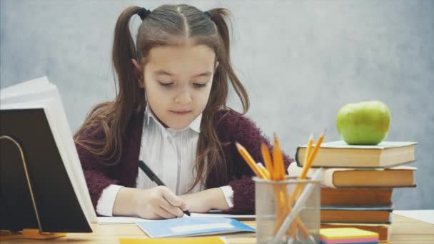 Intelligens iskoláslány egy szürke háttér. Ez idő alatt az asztalnál ül. Óvatosan írja házi feladat. — Stock videók