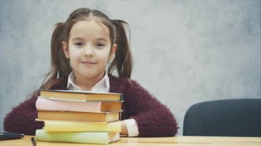 Masada bir kız öğrenci oturuyor. Bu dans sırasında kameraya bakıyor. Bir yığın kitaba sarıldıktan sonra başını kaldırdı. Gri arka planda.