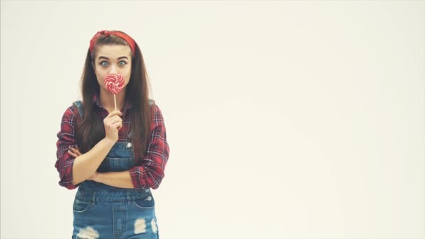 Reizendes Mädchen, das seinen Mund hinter dem roten herzförmigen Lolli-Pop versteckt, dann lächelt und die Augenbraue hochzieht. — Stockvideo