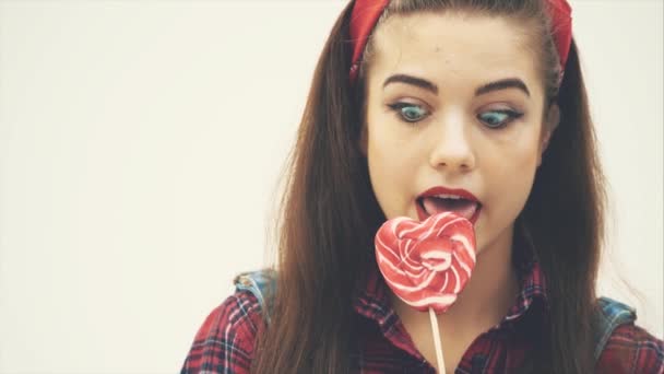 Bella ragazza pinup leccare rosso a forma di cuore lecca-lecca, espressione del viso soddisfatto . — Video Stock