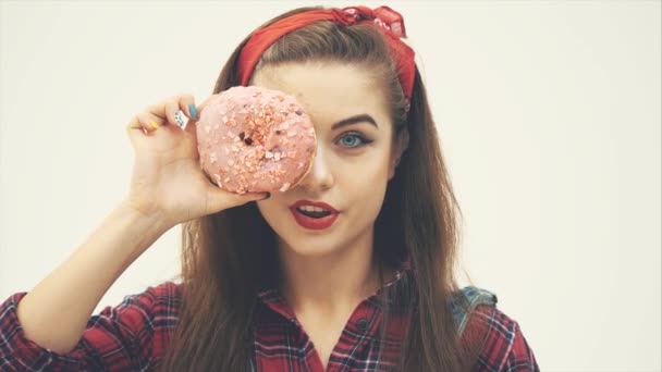 Linda garota pinup está escondendo seu olho atrás de um grande donut rosa, piscando, levantando a sobrancelha, sorrindo . — Vídeo de Stock