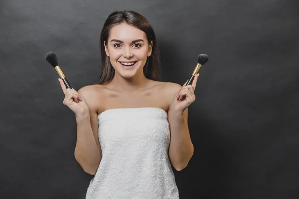 Happy smiling brunette girl in white towel holding make up brushes. She holds the brush in both hands. — Stock Photo, Image