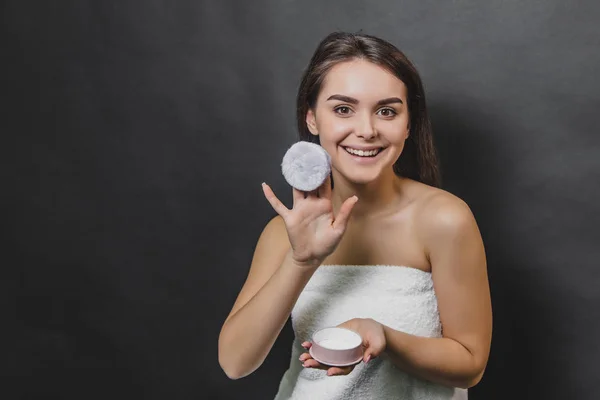 Portrait of young woman wrapped in towel standing isolated over black background, holding powder puff. — Stock Photo, Image