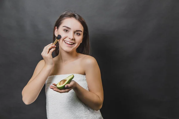 Hermosa joven se encuentra sobre un fondo negro. Durante este tiempo sostenga un cepillo de aguacate y maquillaje. Tiene una piel bastante limpia . — Foto de Stock