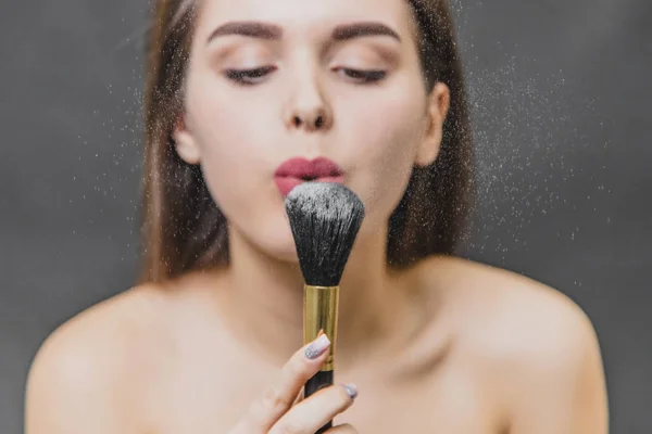 Gadis muda yang cantik memegang sikat untuk makeup di tangannya. Ini meniup dari bubuk untuk makeup dan melihatnya. Aplikasi makeup dalam close-up . — Stok Foto