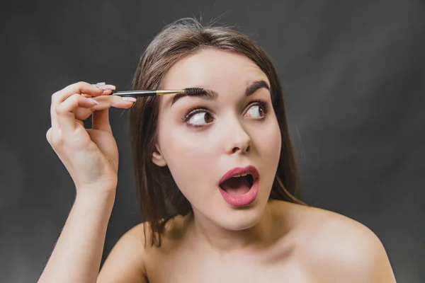 Belle jeune fille se tient sur un fond noir. Pendant ce temps, en regardant ailleurs à l'aide d'une brosse, fait un maquillage des sourcils . — Photo