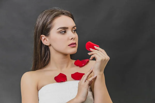 Beautiful young girl stands on a black background. During this time in the spa salon after the procedures on the body has a petal of red rose.
