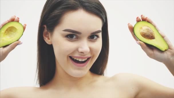 Young pretty girl standing on a white background. The concept of living products. Close-up portrait of a beautiful young woman with long healthy hair holding an green avocado near her cheeks. Copy — Stock Video