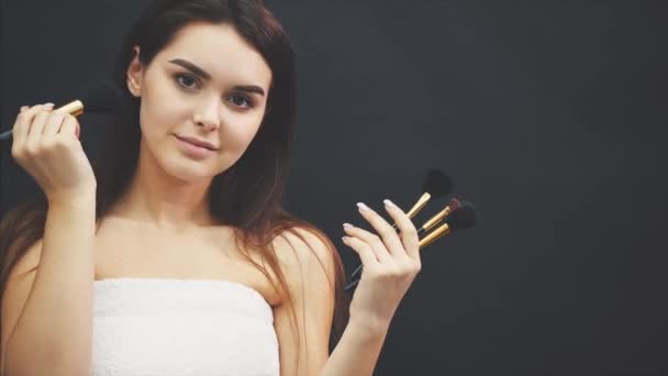 Joven linda chica de pie sobre un fondo negro. Al mismo tiempo sosteniendo un pincel en su mano y llevándolo a cabo en la cara. Sonriendo muestra un beso de aire . — Vídeos de Stock