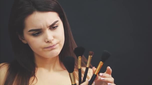 Ung ganska söt flicka stående på en svart bakgrund. Samtidigt håller den i hand borstar för make-up att välja den som behövs. Har en ganska ren hud. — Stockvideo