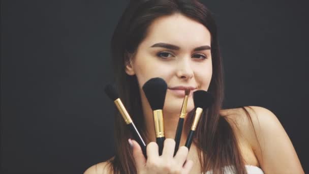 Belleza modelo chica, artista de maquillaje sosteniendo conjunto de maquillaje cepillos y sonriendo. Hermosa joven morena con piel perfecta y maquillaje desnudo. Un primer plano perfecto de la piel. Maquillaje de contorno facial . — Vídeos de Stock