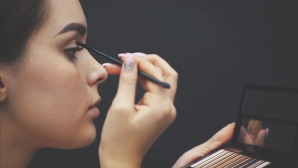 Közeli fel egy gyönyörű arca egy fiatal lány kap egy átalakítása. Nő alkalmazó szemhéjfesték a szemöldökét a kefe. — Stock videók