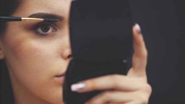 Menina bonita fica em um fundo preto. Durante isso, olhando um espelho e usando um pincel faz uma maquiagem sobrancelha . — Vídeo de Stock
