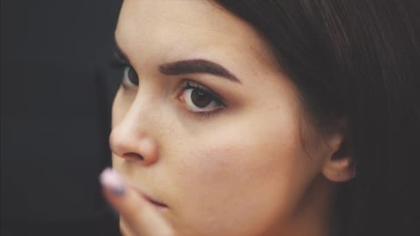 Schöne junge Mädchen steht auf einem schwarzen Hintergrund. Dabei macht der Blick in den Spiegel und die Verwendung eines Pinsels ein Augenbrauen-Make-up.. — Stockvideo