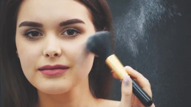 Ein junges Mädchen streicht sich mit einem Schminkpinsel ein Puder über das Gesicht. Make-up-Anwendung in Nahaufnahme. isoliert auf schwarzem Hintergrund. das Mädchen hat eine saubere Haut und schöne lange Haare. — Stockvideo