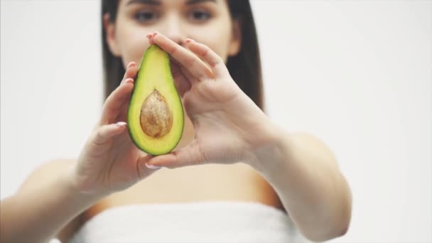 Jong aantrekkelijk meisje staat in een studio op een zwarte achtergrond. Gedurende deze tijd kijkt hij naar de camera en verhoogt hij de avocado op de meisjes wazige achtergrond. Heeft een schone huid en lang mooi haar. — Stockvideo