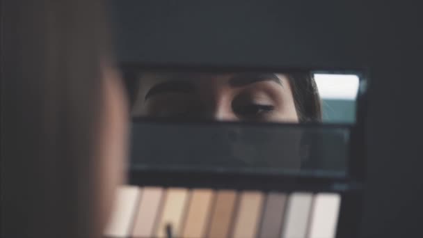 Close-up van het gezicht van een jonge vrouw krijgen een make-up. Een mooie vrouw die een oogschaduw op haar wenkbrauwen aanbrengt, is een borstel. Mooi meisje op zoek in de spiegel voor schaduwen. Close-up. — Stockvideo