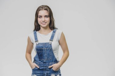 Genç güzel öğrenci kız beyaz bir arka plan üzerinde duruyor. Kot takım elbise ve beyaz bir tişört giymiş. Ellerini cebine sokarak, kameraya gülümseyerek bakıyor..