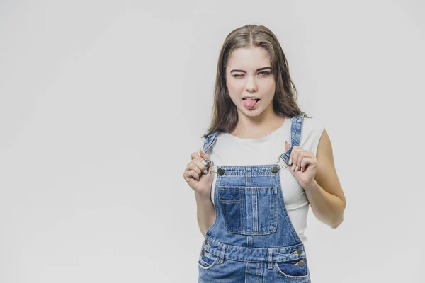 Junges hübsches Studentenmädchen, das auf weißem Hintergrund steht. bekleidet mit einem Jeansanzug und einem weißen T-Shirt. Er hält seine Hände an der Zange aus der Kombination und schaut in die Kamera, schließt ein Auge. — Stockfoto