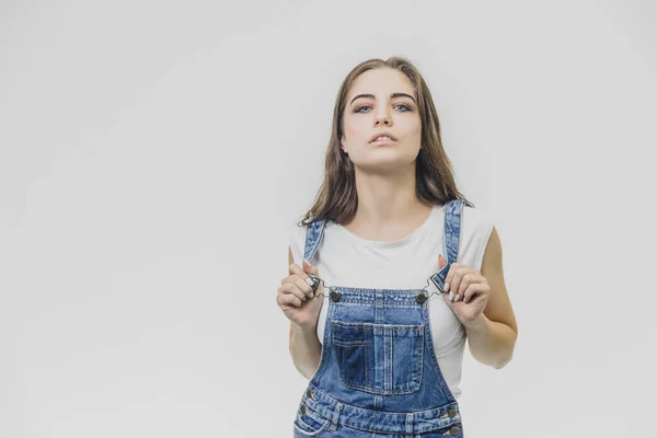 Jovem menina estudante bonita de pé sobre um fundo branco. Vestida com um fato de ganga e uma t-shirt branca. Segurando as mãos no alicate da combinação, ele olha para a câmera, levantando a cabeça — Fotografia de Stock