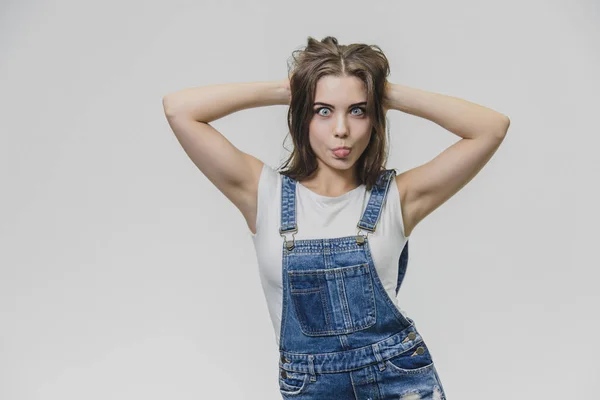 Una adolescente con placer una mirada atractiva. Lleva un mono de mezclilla de moda y una camisa blanca. Pon las manos sobre la cabeza y muestra la lengua. El concepto de personas, estilo y moda —  Fotos de Stock