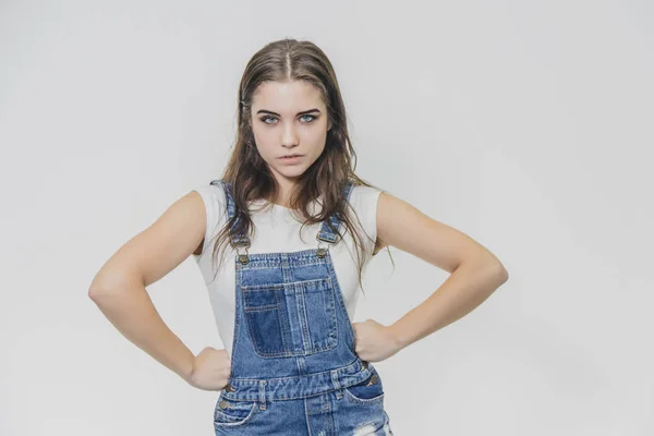 Uma jovem menina bonita fica em um fundo cinza. Ele está vestido com um terno de ganga e uma camiseta branca. Durante isso, ele segura seus braços na cintura e olha para a câmera . — Fotografia de Stock