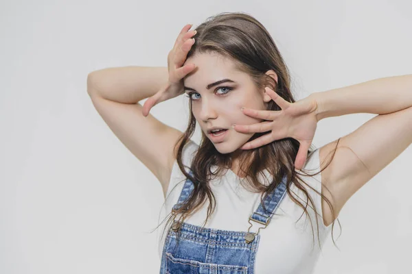 Una chica joven y hermosa está de pie sobre un fondo gris. Está vestido con un traje de mezclilla y una camiseta blanca. Durante esto, él sostiene sus brazos en su cintura y mira a la cámara . —  Fotos de Stock