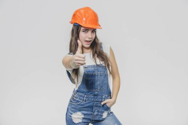 Beyaz bir arka plan üzerinde duran genç güzel kız. Bu süre zarfında beyaz bir t-shirt ve kot tulum giyiyor. Turuncu bir kask giymiş.