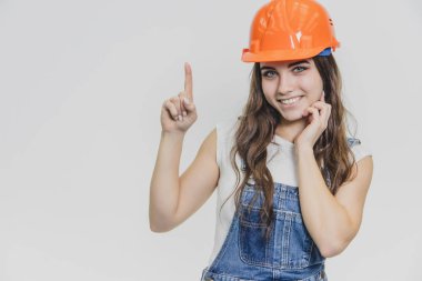 Genç ve güzel bir kız gri bir arka plan üzerinde duruyor. Kafasına turuncu bir kask giymiş. Bu esnada kameraya bakar ve işaret parmağını yukarı gösterir..