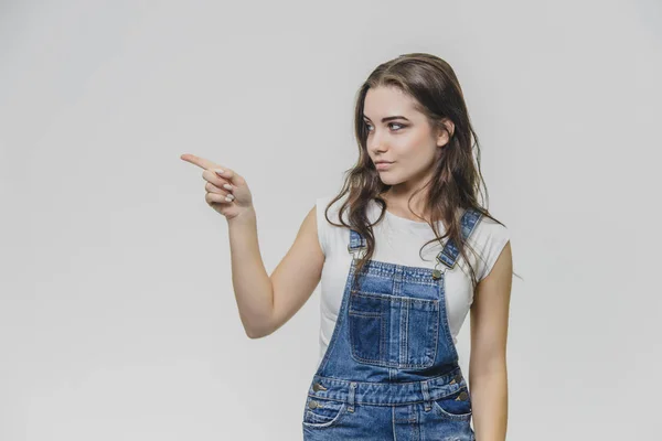 Una joven estudiante atractiva muestra su dedo índice a un lado. Intenta concentrarse y mira a un lado. Vestido con camiseta blanca y overoles de mezclilla. Aislado sobre fondo blanco . —  Fotos de Stock