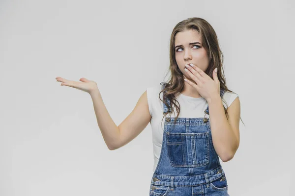 Young student girl standing on a white background. Dressed in white t-shirt and denim overalls. During this time, putting one hand to one side, the other hand shields his mouth and looks at the camera — Stock Photo, Image