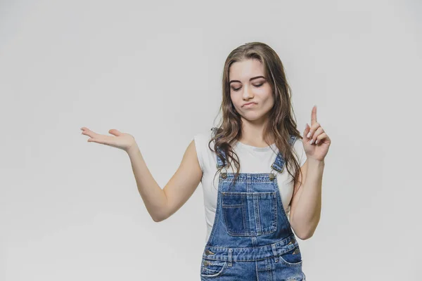 Young student girl standing on a white background. Dressed in white t-shirt and denim overalls. During this, putting one hand to one side, the other hand points with the index finger up and closing — Stock Photo, Image