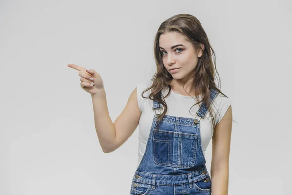 Una joven estudiante atractiva muestra su dedo índice a un lado. Intenta concentrarse y mira a la cámara. Vestido con camiseta blanca y overoles de mezclilla. Aislado sobre fondo blanco . — Foto de Stock