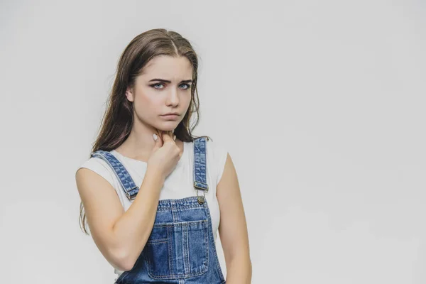 Uma jovem e tensa mulher bonita segura os braços ao redor do pescoço, sente emoções doentias. Sofre de dor após o ar frio. Usa uma t-shirt branca, macacão, posa dentro de casa . — Fotografia de Stock