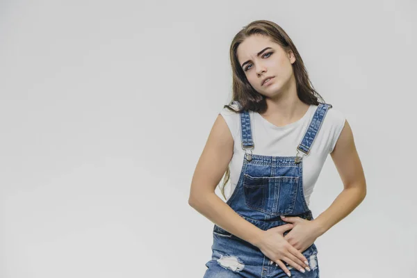 Uma jovem e tensa mulher bonita segura os braços em seu estômago, sente emoções dolorosas. Ela sofre de dor após o trabalho duro. Uma t-shirt branca bonita, macacão, poses dentro de casa . — Fotografia de Stock