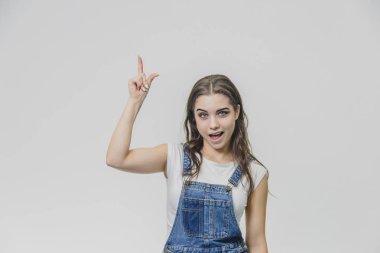 Genç çekici öğrenci kız onu işaret parmağı kadar gösterir. Konsantre olmaya çalışır ve kameraya bakar. Beyaz t-shirt ve kot tulum giymiş. Beyaz arka plan üzerinde yalıtılmış.