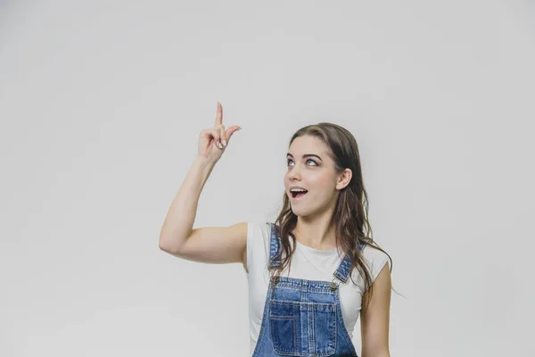 Jovem menina estudante atraente mostra seu dedo indicador para cima. Tenta concentrar-se e olha para a câmara. Vestida com t-shirt branca e macacão de ganga. Isolado sobre fundo branco . — Fotografia de Stock