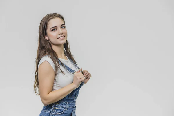 A cheerful cute teen girl holds his hands on the pliers. Has a surprised facial expression and looks at the camera with a sincere smile. Dressed in white t-shirt and denim overalls. — Stock Photo, Image