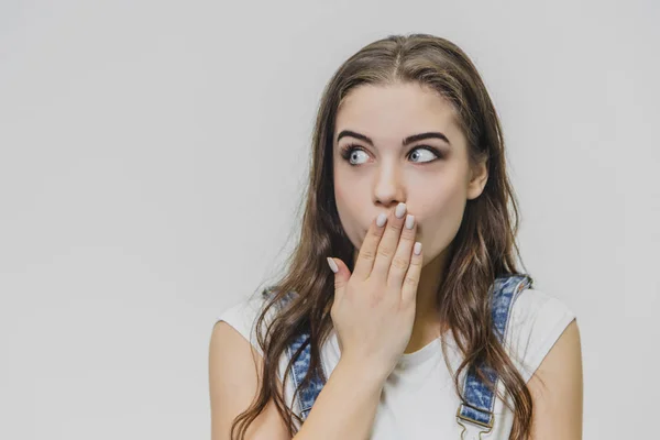 Ritratto di una bella donna con espressione stupita del viso. Indossa una tuta di jeans e una maglietta bianca. Isolato su sfondo bianco. La ragazza guarda di lato mettendo la mano su di lei — Foto Stock