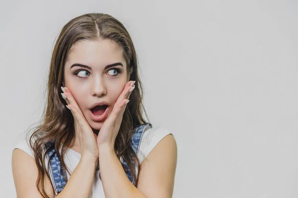 Retrato horizontal de uma bela mulher com expressão espantada do rosto. Usa um macacão de ganga e uma t-shirt branca. Isolado sobre fundo branco . — Fotografia de Stock