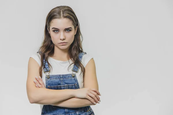 La joven y hermosa niña estaba de pie sobre un fondo blanco. Vestido con un mono de mezclilla y una camiseta blanca. Durante este tiempo, la colocación de una mano en la mano mira cuidadosamente a la cámara . —  Fotos de Stock