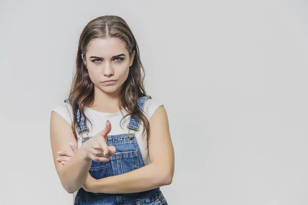 Young pretty girl standing on a white background. Dressed in a denim overalls and a white T-shirt. During this time, place your index finger forward. — Stock Photo, Image