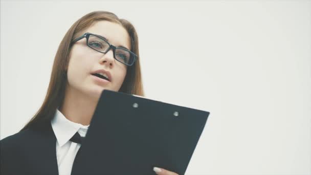 La mujer de negocios está sobre un fondo blanco. Al mismo tiempo, sostiene una carpeta negra con documentos que la abrazan cuidadosamente mirando a la cámara. Sonrisas fuertes . — Vídeos de Stock