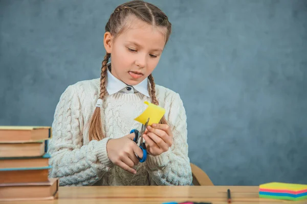 La bambina sta tagliando un adesivo giallo con le forbici mentre è seduta alla scrivania, isolata su sfondo grigio . — Foto Stock