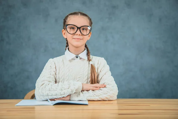 Linda, encantadora chica sonríe con las manos dobladas y lleva gafas mientras está sentado en el escritorio y se concentra . — Foto de Stock