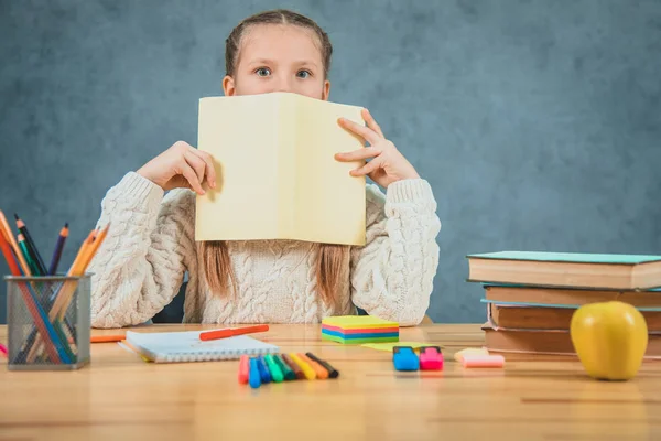 Charmig Schoolgirl ser ut gult pappersark sitter vid skrivbordet isolerad på grå bakgrund. — Stockfoto