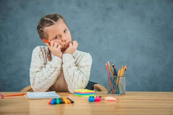 Uttråkad Schoolgirl sätter huvudet nedåt och underställer huvudet med händerna. Liten flicka komprimerar röd penna på grå bakgrund. — Stockfoto