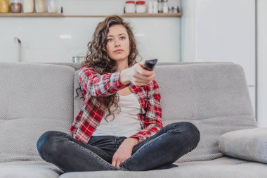 Güzel bir kadın TV izliyor ve kanepede oturuyor ve elinde uzaktan kumandayı tutuyor. Bir gömlek bir esmer uzun saçlı TV bakıyor.