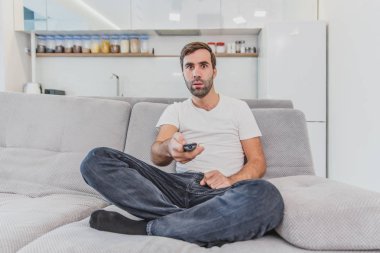 Uzaktan kumandayı tutan güzel, komik bir genç adam. Bu sırasında, TV evde kanepede otururken izliyor.