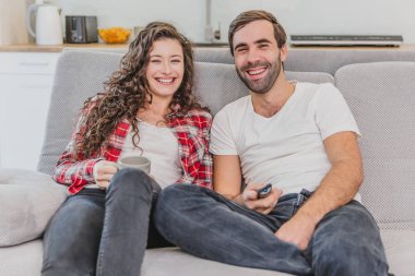 Güzel komik genç adam bir uzaktan kumanda tutar, kahve karısı fincan. Bu esnada kameraya bakıp gülümsüyorlar. Evde kanepede oturmak.